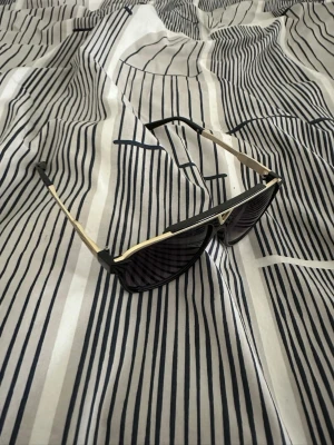 Svarta LV solglasögon - Snygga solglasögon från LV med svart båge och guldiga detaljer. Rektangulär form och mörka glas. Märkeslogga på skalmen för extra lyxig känsla. Perfekta för dig som vill sticka ut med en exklusiv accessoar.