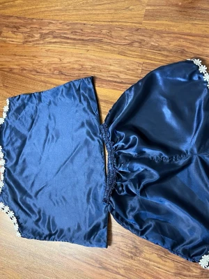 Mörkblått satinsovset med spets - Sött tvådelat sovset i mörkblå satin med vita blommiga spetsdetaljer längs kanterna. Setet består av ett linne och ett par shorts med resår i midjan. Materialet är glansigt och mjukt, perfekt för en lyxig känsla hemma.