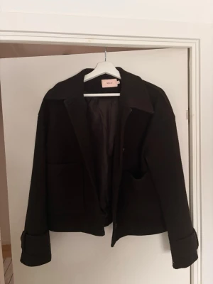 Svart kappa från Nelly - Kort snygg svart kappa från Nelly i storlek xs. Modellen har klassisk krage, raka ärmar med knappdetalj vid mudden och två stora fickor framtill. Kan stängad med knappar.  Passar bra för vår och sommar.