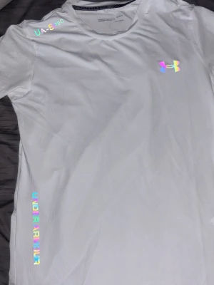 Vit träningströja från Under Armour - Snygg vit träningströja från Under Armour med holografiska detaljer och logga på bröstet. T-shirten har rund halsringning och korta ärmar, tillverkad i ett lätt och snabbtorkande material som passar perfekt till träning.