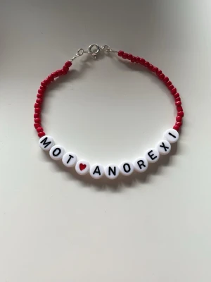Pärlhalsband med text mot anorexi - Unikt armband med röda små pärlor och vita bokstavspärlor som stavar 'MOT ANOREXI' med ett rött hjärta. Armbandet har ett enkelt metallås och är handgjort i plast. Perfekt för dig som vill bära ett statement.