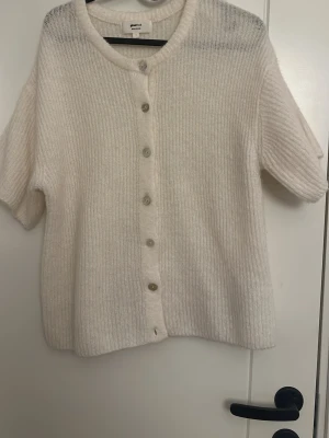 Vit kofta från Gina Tricot - Mysig vit kofta från Gina Tricot i ullblandning. Koftan har rund halsringning, korta ärmar och knäppning med genomskinliga knappar framtill. Ribbstickad struktur ger en trendig och avslappnad look.