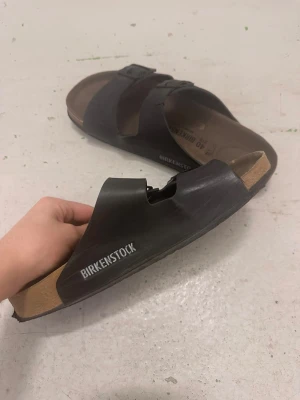 Svarta Birkenstock sandaler med spännen - Klassiska svarta Birkenstock sandaler med två breda remmar och justerbara metallspännen. Fotbädden är i kork och mocka för extra komfort. Perfekta för en avslappnad och trendig stil. Birkenstock-logga på sidan.