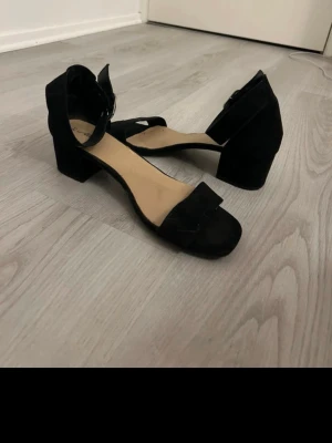 Svarta sandaler med blockklack - Stilrena svarta sandaler med bred rem över tårna och justerbart spänne runt ankeln. Skorna har en bekväm blockklack och öppen tå. Materialet ser ut att vara mockaimitation och innersulan är ljusbeige. Perfekta för dig som gillar enkel men trendig stil.