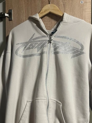 Beige hoodie från FSR med blingtryck - Säljer en oversized beige hoodie från FSR i storlek S. Hoodien har dragkedja, fickor framtill och ett stort silvrigt blingtryck över bröstet. Tillverkad i mjukt material och har en klassisk huva. Perfekt för dig som gillar streetwear vibes.