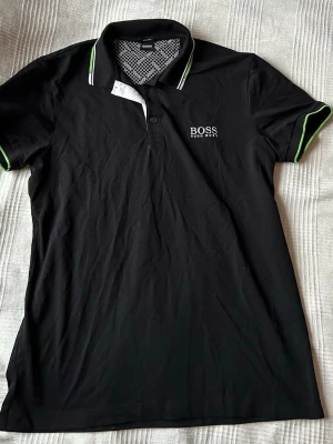 Svart pikétröja från Hugo Boss - Snygg svart pikétröja från Hugo Boss med vit logga broderad på bröstet. Tröjan har krage med vita och gröna detaljer samt gröna ränder på ärmsluten. Klassisk passform och knappslå framtill. Perfekt för en stilren look.