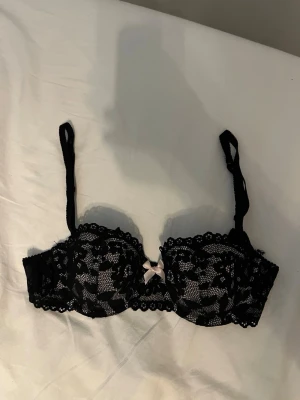 Svart spets-bh med rosett - Snygg svart bh med spetsdetaljer och en liten ljus rosett framtill. Bh:n har justerbara axelband och bygel för extra stöd. Spetsen har ett blommigt mönster och kanten är vågig för en feminin look.