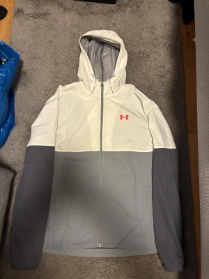 Under Armour tvåfärgad vindjacka - Snygg vindjacka från Under Armour med vit överdel och grå nederdel samt ärmar. Jackan har en huva, hel dragkedja och meshfoder på insidan för extra ventilation. Under Armour-loggan i rosa på bröstet. Perfekt för att hålla vinden borta under träning eller ute. Använd 3 gånger! Hör av er vid byten också