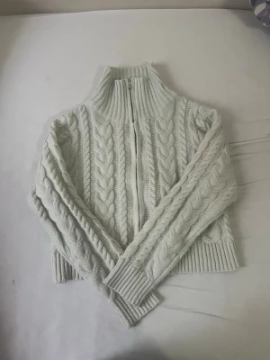 Ljusgrå kabelstickad ziptröja - Säljer en ljusgrå kabelstickad tröja med dragkedja framtill och hög krage. Tröjan har långa ärmar och breda ribbade muddar vid ärmslut och nederkant. Perfekt för lager på lager och riktigt mysig att ha på sig.