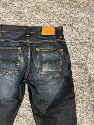 Mörkblå jeans från Nudie Jeans - Snygga mörkblå jeans från Nudie Jeans med klassisk femficksmodell och kontrastsömmar. Jeansen har slitningar och tvättade detaljer för en cool, använd look. Tillverkade i denim och har riktigt schyssta fades✌️dem är i storlek W30 och L34