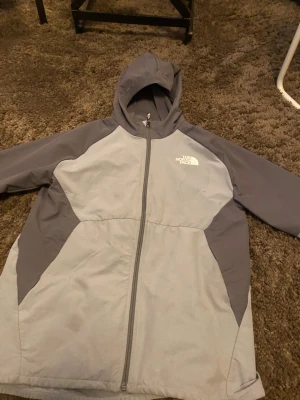 Grå vindjacka från The North Face - Snygg grå vindjacka från The North Face med två nyanser av grått, hel dragkedja och huva. Jackan har logga på bröstet och är tillverkad i ett lätt, vindtätt material. Perfekt för blåsiga dagar och enkel att bära med sig. Skriv för mer information/ bilder 