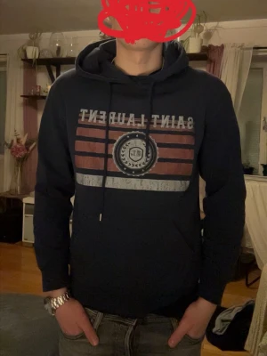 Mörkblå Saint Laurent hoodie - Snygg mörkblå hoodie från Saint Laurent med tryck framtill i vitt och rött samt ett emblem i mitten. Klassisk huva med dragsko och känguruficka. Perfekt för dig som vill ha en stilren och trendig look med streetwear-vibe.