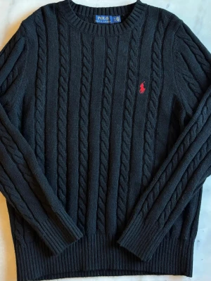 Svart kabelstickad tröja Polo Ralph Lauren -  Svart tröja från Polo Ralph Lauren med rund halsringning och röd broderad logga på bröstet. Tröjan har långa ärmar och ribbade muddar vid ärmslut och nederkant. Klassisk design i mjukt material.