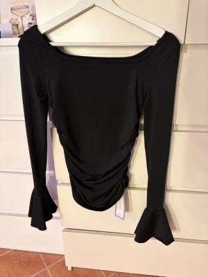 Svart långärmad topp med volangärm - Trendig svart topp med långa ärmar och volangdetaljer vid ärmsluten. Toppen har en tight passform och rynkade sömmar längs sidorna som ger en snygg siluett. Tillverkad i mjukt, stretchigt material som känns bekvämt mot huden.