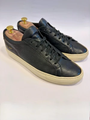  Common Projects Achilles Low EU40 Grained Leather - Stilrena svarta sneakers från Common Projects i mjukt premiumläder med diskret guldfärgad numrering på sidan. Klassisk låg modell med vita gummisulor och svarta skosnören. Perfekta för en clean och modern look.