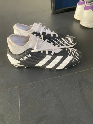 Adidas Predator svartvita fotbollsskor - Säljer ett par Adidas Predator fotbollsskor i svart och vitt med klassiska tre ränder på sidan. Skorna har snörning, låg profil och mönstrad ovandel för bättre bollkontroll. Tillverkade i syntetmaterial och designade för spel på gräsplan.  