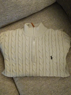 Ralph Lauren Kabelstickad Tröja med Dragkedja - Ny med prislapp - Säljer en snygg och klassisk kabelstickad tröja från Ralph Lauren, modell Polo. Tröjan är i ljusgrå färg och har en praktisk dragkedja vid halsen samt en stilren krage. Perfekt för både vardagsbruk och mer uppklädda tillfällen. Färg: Ljusgrå Material: 100% Bomull  Storlek: Xl (men passar perfekt till er som har L) En tidlös och lyxig tröja som håller dig varm och snygg hela säsongen. Missa inte chansen att äga en äkta Ralph Lauren!