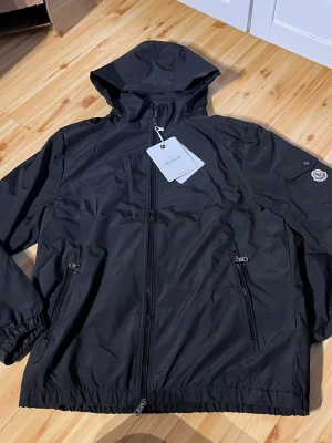 Svart vindjacka från Moncler - Snygg svart vindjacka från Moncler med huva, dragkedja framtill och två fickor med dragkedja. Jackan har elastiska muddar vid ärmslut och nederkant samt Moncler-logga på ärmen. Tillverkad i lätt och slitstarkt material, perfekt för blåsiga dagar.