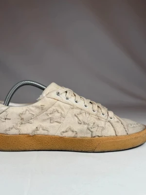 Saint Laurent beige sneakers med mönster - Unika beige sneakers från Saint Laurent med slitna detaljer och broderade stjärnor på canvasmaterial. Skorna har klassisk snörning och en kontrasterande brun gummisula som ger en cool vintagekänsla. Perfekta för dig som gillar streetstyle och exklusiva märken. Storlek 42 men passar 43