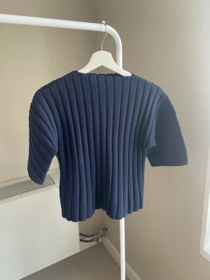 Mörkblå ribbstickad topp - Säljer en mörkblå ribbstickad topp med korta ärmar och rund hals. Tröjan har en cool struktur med breda vertikala ribbor som ger ett modernt intryck. Perfekt att styla med jeans eller kjol för en trendig look.