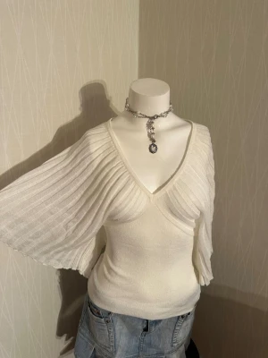 Vit ribbad stickad topp med v-ringning - Säljer en vit topp med bred v-ringning och ribbade, vida ärmar. Toppen har en figurnära passform och är tillverkad i ett mjukt, stretchigt material som känns bekvämt mot huden. Perfekt för dig som gillar en stilren men ändå unik look.