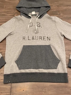 Grå hoodie från Ralph Lauren - Snygg grå hoodie från Ralph Lauren Denim & Supply med mörkare grå huva och magficka. Tryck på bröstet med texten 'U.S. R. LAUREN' och ett litet sköldmärke. Mjuk bomullsblandning och dragsko i huvan. Perfekt för en avslappnad streetwear-look. ❗️OBS inte äkta. Passa dig mellan 175-185. 