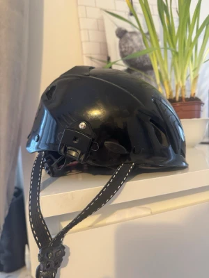 Svart arbets hjälm med justerbar rem - Säljer en svart taktisk hjälm med flera ventilationshål och justerbar hakrem med vit söm. Hjälmen har fästen för tillbehör och är tillverkad i hårdplast. Perfekt för airsoft, paintball eller liknande aktiviteter.