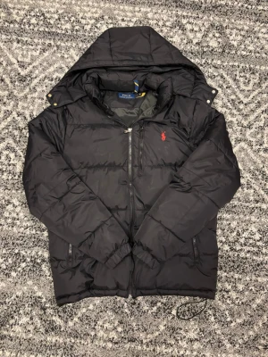 Svart pufferjacka från Polo Ralph Lauren - Snygg svart pufferjacka från Polo Ralph Lauren med huva och dragkedja framtill. Jackan har det röda Polo-logotrycket på bröstet och sidofickor med dragkedja. Jackan är i storlek S men kan även passa i storlek M. Påsen kommer med!! Hör av er vid eventuella frågor eller funderingar!!