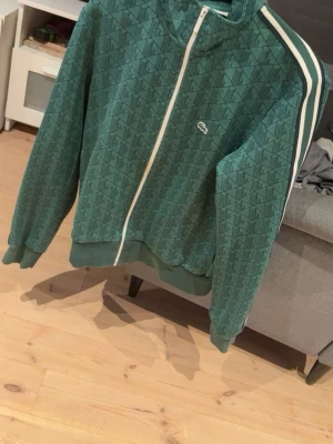 Grön zip hoodie från Lacoste - Säljer en grön zip hoodie från Lacoste med diskret mönster och vit dragkedja. Klassisk krokodil-logga på bröstet och vita ränder längs ena ärmen. Ribbstickade muddar och krage. Perfekt för en sportig och avslappnad stil.