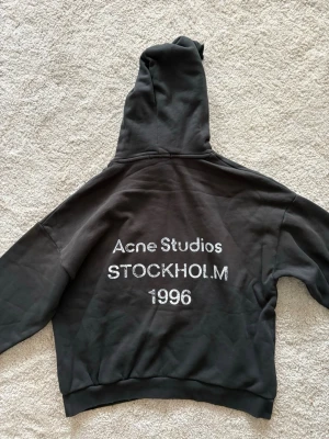 Acne studios ”Stockholm” hoodie - Säljer en svart hoodie från Acne Studios med tryck på ryggen där det står 'Acne Studios STOCKHOLM 1996'. Hoodien har en stor huva och ribbade muddar. Materialet är mjuk bomull, perfekt för en avslappnad och trendig look.lappen är inte kvar!! Äkta! 