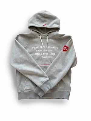Peak Performance Åre Hoodie - Otroligt fet grå hoodie från Peak Performance med vit text framtill och röd detalj vid fickan. Lappen borta på insidan men sitter som en M. Förutom det är den i perfekt skick.