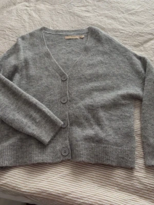 Grå kofta från Culture - Mysig grå kofta från Culture med v-ringning och fyra stora knappar framtill. Koftan är stickad i ett mjukt material och har ribbade muddar vid ärmslut och nederkant. Använd fåtalgånger. Ny pris 1200kr