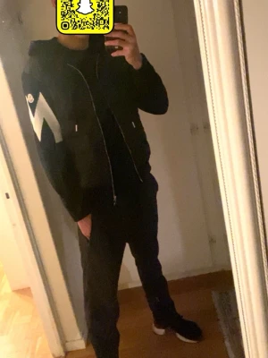 Svart Moncler hoodie med dragkedja - Säljer en svart hoodie från Moncler med vadderad framsida och stickade ärmar. Dragkedja framtill, huva med snören och en stor vit Moncler-logga på ena ärmen. Snygg och modern design som passar perfekt till streetwear-stilen.