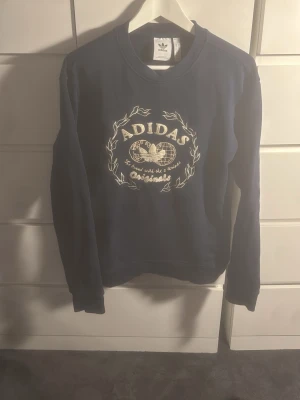 Mörkblå sweatshirt från Adidas - Mörkblå sweatshirt från Adidas Originals med broderad vit logga och text på bröstet. Tröjan har rund halsringning och långa ärmar. Perfekt för en avslappnad streetwear-look.