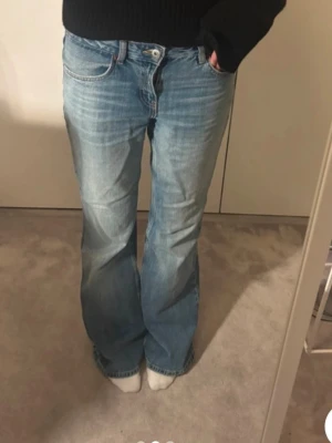 Blå bootcut jeans med ljus tvätt - Säljer ett par blå bootcut jeans med ljus tvätt och klassisk femficksdesign. Jeansen har normal passform och är tillverkade i jeansmaterial med snygga slitningar framtill. Perfekt för dig som gillar retrovibbar och vill ha ett par sköna baggy jeans med lite vidare ben