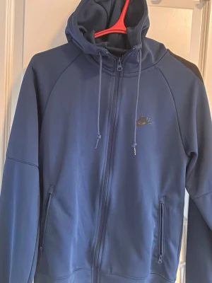 Blå zip hoodie från Nike - Snygg blå hoodie från Nike med dragkedja framtill och två fickor med dragkedja. Hoodien har justerbar huva med snören och en diskret Nike-logga i silver på bröstet. Tillverkad i mjukt och bekvämt material, perfekt för en avslappnad stil.