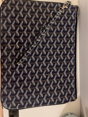 G O Y A R D väska  - Säljer en mörkblå kuvertväska från Gyard med det klassiska vita mönstret och silverfärgad dragkedja. Väskan har en avtagbar handledsrem och insidan är fodrad i gult tyg med en liten ficka. Perfekt för att bära det viktigaste med stil.