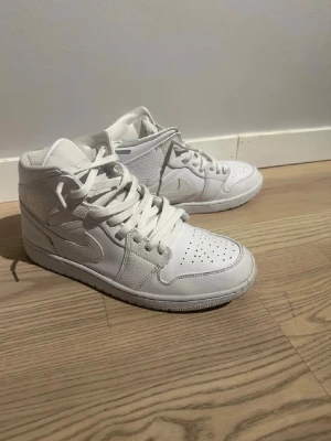 Vita Nike Air Jordan 1 Mid sneakers - Säljer ett par helvita Nike Air Jordan 1 Mid sneakers med klassisk siluett. Skorna har perforerad tå, vita skosnören och ikoniska swoosh-loggan på sidan. Tillverkade i läder och textil för en clean och stilren look.