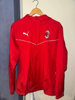 Röd AC Milan vindjacka från Puma - Snygg röd vindjacka från Puma med AC Milan-logga på bröstet och vit Puma-logga. Jackan har huva, dragkedja framtill och elastiska muddar. Perfekt för träning eller när du vill representera ditt lag.
