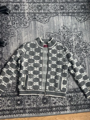 Gråvit stickad kofta från Gucci - Säljer en grå stickad kofta från Gucci med vit GG-monogram över hela plagget. Koftan har dragkedja framtill, ribbade muddar och två fickor. Perfekt för dig som gillar exklusiv streetwear och vill sticka ut med en ikonisk design.