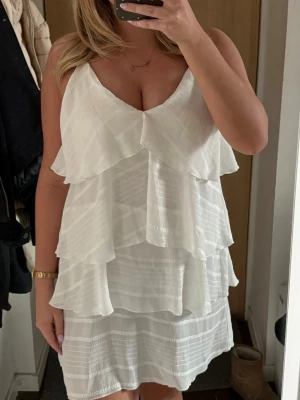 Vit omlottklänning med volanger - Säljer en vit kortklänning med omlott-design och flera lager volanger. Klänningen har v-ringad hals och är ärmlös, perfekt för sommarens alla tillfällen. Materialet är lätt och luftigt, vilket gör den bekväm att bära.