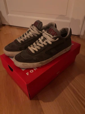 Gråa Diesel sneakers med vita detaljer - Snygga gråa sneakers från Diesel med vita snören och vit sula. Skorna har en klassisk låg profil och Diesel-logga i rött på plösen. Materialet ser ut att vara mocka och textil, vilket ger en cool och avslappnad vibe. Sitter större på foten. Som en 40,5
