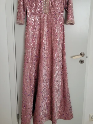 Glittrig rosa långklänning med paljetter - Elegant långklänning i rosa med glittriga paljetter. Klänningen har trekvartsärm och dekorativa detaljer längs halsen och ärmsluten. Perfekt för speciella tillfällen när du vill sticka ut.