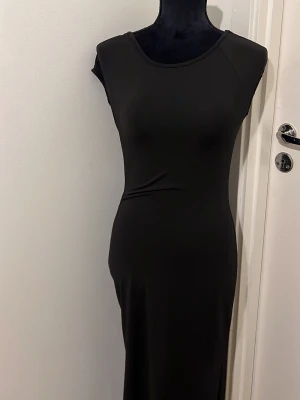Svart långklänning med rund hals - En stilren svart långklänning med rund halsringning och ärmlös design. Klänningen har en figurnära passform och är tillverkad i ett mjukt, stretchigt material som ger en smickrande siluett. Perfekt för dig som gillar minimalistisk och tidlös stil.