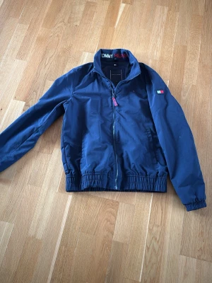 Marinblå vindjacka Tommy Hilfiger - Snygg marinblå vindjacka från Tommy Hilfiger med dragkedja framtill. i nacken är det en logga vit och röd. storlek 176.  Jackan är använd men inget som direkt syns.