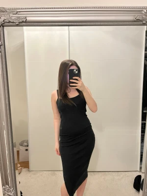 Svart asymmetrisk klänning - Säljer en svart, ärmlös klänning med asymmetrisk nederkant och slits. Klänningen har en smal passform och enkel, stilren design. Perfekt för dig som gillar minimalistiskt mode och vill ha något som sticker ut med sin unika skärning.