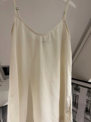 Krämvitt linne med smala axelband i siden - Krämvitt linne med smala axelband i siden. Sött krämvitt linne med tunna, justerbara axelband och diskret spetskant vid urringningen. linnet har en rak och luftig passform även lite tajt, perfekt för mysiga kvällar hemma. Materialet ser ut att vara mjukt och följsamt.