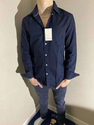 Mörkblå skjorta från these glory days - Helt ny Stilren mörkblå skjorta från these glory days med klassisk krage och vita knappar framtill. Skjortan har lång ärm och är tillverkad i ett mjukt bomullstyg som känns bekvämt mot huden. Perfekt för en clean och snygg look. Slim fit så sitter sjukt bra. Nypris 1099kr