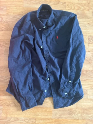 Marinblå skjorta från Ralph Lauren - Klassisk marinblå skjorta från Ralph Lauren med röd broderad logga på bröstet. Skjortan har knappar framtill och på manschetterna samt en tidlös passform. Perfekt för dig som gillar stilrena och ikoniska plagg.