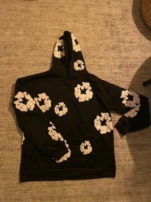 Svart denim tears hoodie - Säljer en svart hoodie med stora vita blommönster över hela tröjan och huvan. Hoodien har klassisk passform, lång ärm och en stor ficka framtill. Perfekt för dig som gillar streetwear och vill sticka ut lite extra.